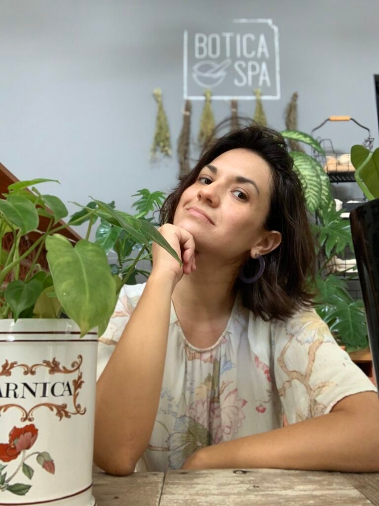 Gina Agsassini, founder of Botica Spa, sitting in a wellness space surrounded by plants and herbal elements.